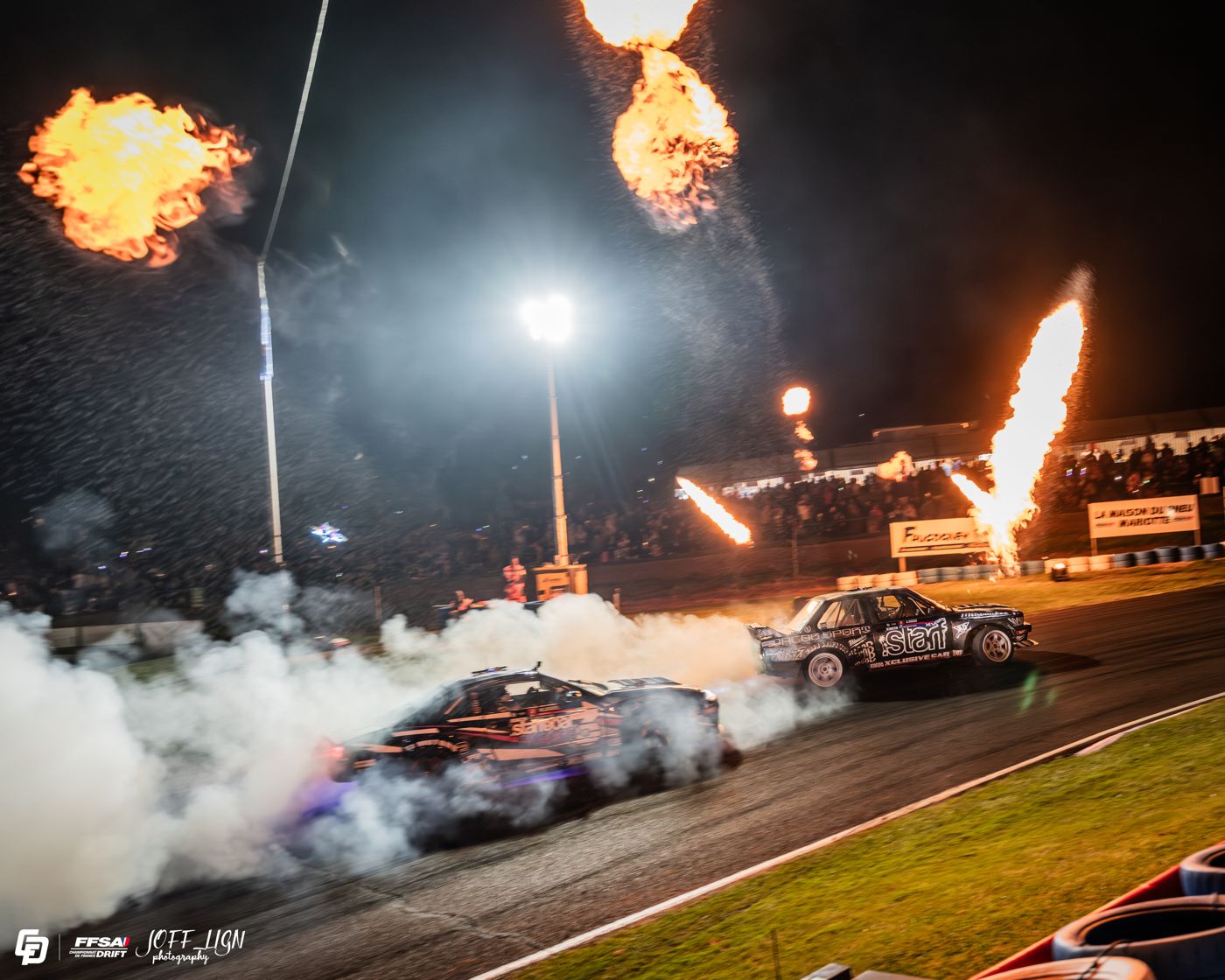 Deux voitures en drift de nuit devant la tribune, sous les flammes de la pyrotechnie